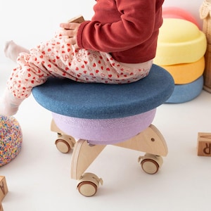 Può includere: Uno sgabello girevole per bambini con seduta blu, sezione centrale lavanda e base in legno con ruote. Lo sgabello ha un design rotondo e a strati, adatto alle aree gioco.