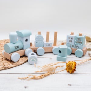 May include: A wooden toy train set in light blue and natural wood tones. The train features the name "Noah" on the engine, and the middle car is 54 cm long. The last car has the time, weight, and date.