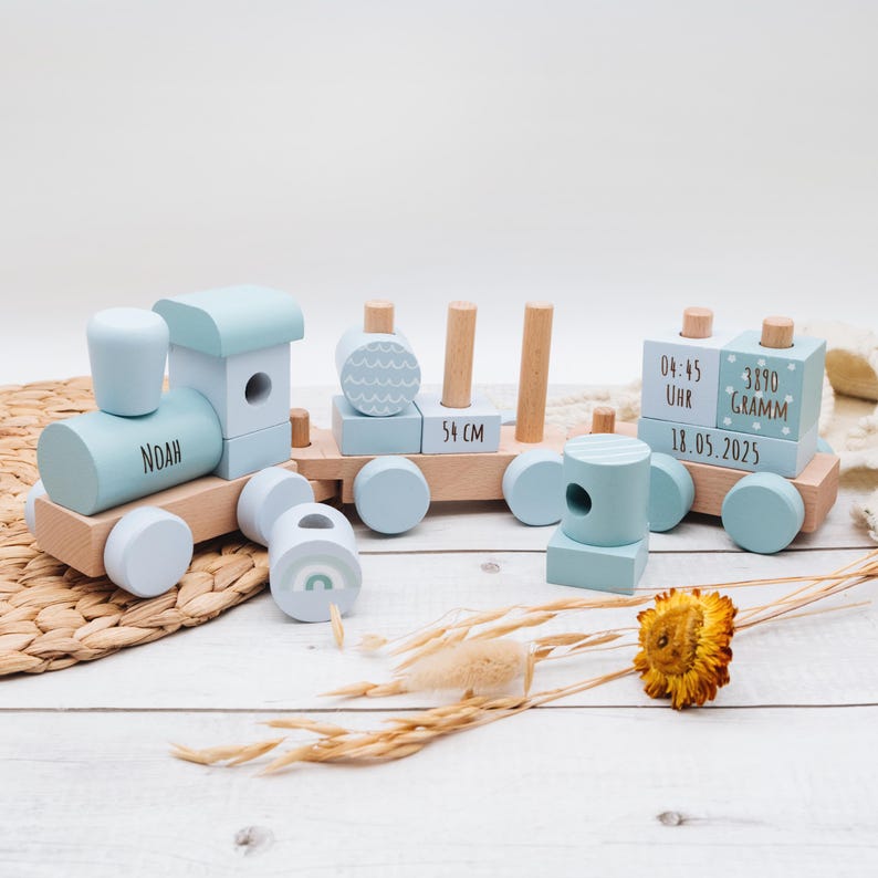 May include: A wooden toy train set in light blue and natural wood tones. The train features the name "Noah" on the engine, and the middle car is 54 cm long. The last car has the time, weight, and date.