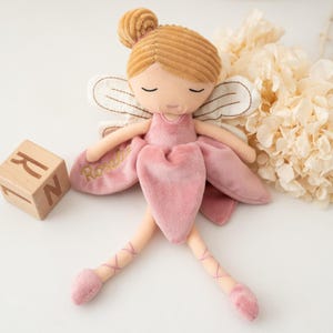 May include: A plush fairy doll in a pink dress with a matching bun and ballet slippers. The doll has closed eyes and embroidered wings. A wooden block with the letter "R" is next to the doll. The name "Rosalie" is embroidered on the dress.