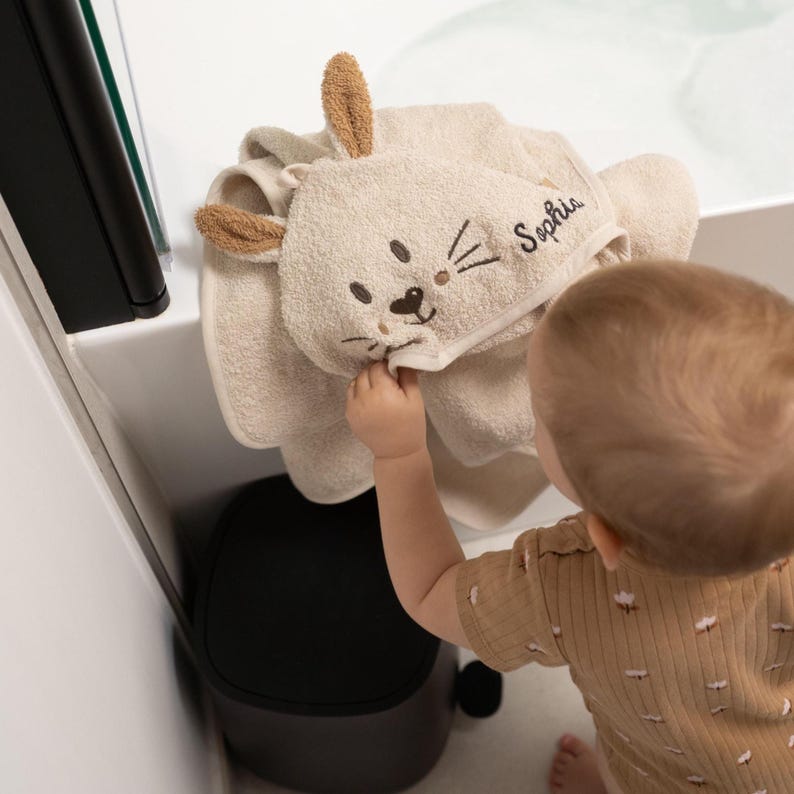 May include: A beige hooded baby towel with a bunny face design. The towel has embroidered details for the eyes, nose, and whiskers. The name "Sophia" is embroidered on the hood. The towel is folded and hanging in a bathroom.