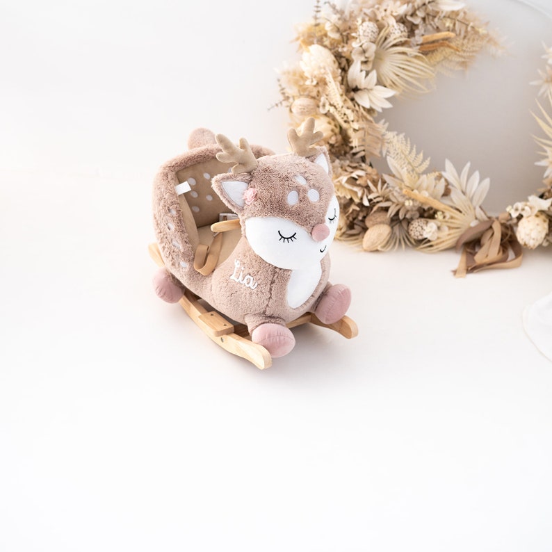 May include: A plush rocking deer toy with a brown and white spotted pattern. The deer has closed eyes and a pink nose. The rocking base is made of wood and the deer has the name "Lia" embroidered on its side.