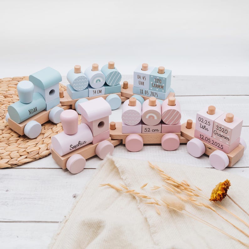 May include: Wooden toy train set in pastel colors. The set includes several train cars in light blue and pink, with details like wheels, smokestacks, and personalized text. Some cars have measurements in centimeters and text with names and dates.