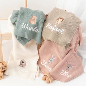 Puede incluir: Una colecci&oacute;n de mantas personalizadas para beb&eacute;s en varios colores, incluyendo verde salvia, beige y rosa. Cada manta presenta un dise&ntilde;o de animal bordado y un nombre en escritura blanca. Las mantas est&aacute;n dobladas y dispuestas en una cesta.