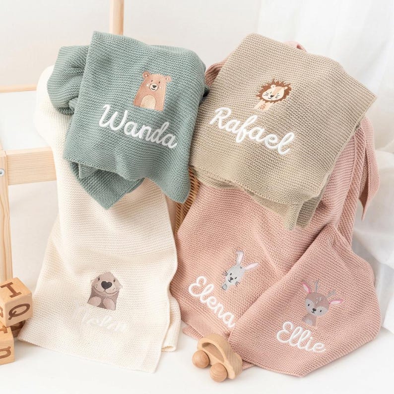 Puede incluir: Una colecci&oacute;n de mantas personalizadas para beb&eacute;s en varios colores, incluyendo verde salvia, beige y rosa. Cada manta presenta un dise&ntilde;o de animal bordado y un nombre en escritura blanca. Las mantas est&aacute;n dobladas y dispuestas en una cesta.