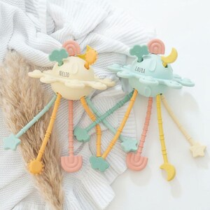 Può includere: Due giocattoli per bambini dai colori pastello, a forma di pianeti con gambe attaccate e forme decorative. Uno è giallo con il nome "NELIO", l'altro è blu con "LAURA". Presentano stelle, nuvole e lune.