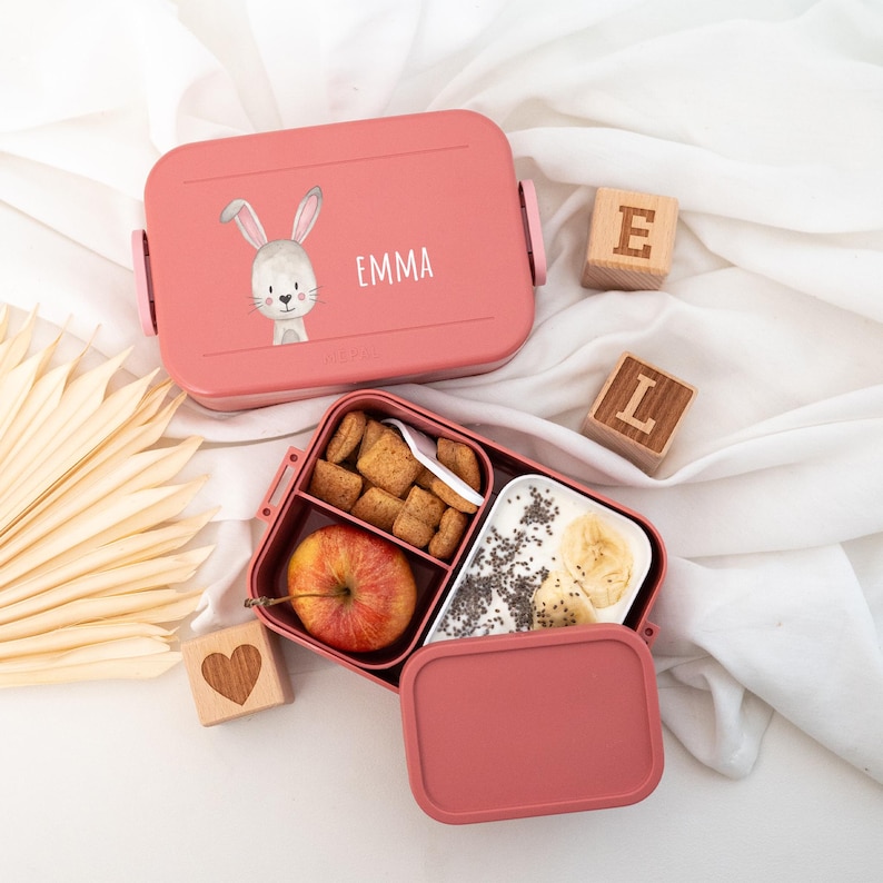 May include: Pink lunchbox with a bunny and the name "Emma" on the lid. The lunchbox is open and contains an apple, cereal, and yogurt with banana and chia seeds.