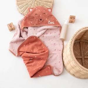 May include: A coral and pink hooded baby towel with a cat face design. The towel has white polka dots and the name "Leni" embroidered on the hood. A matching washcloth is folded next to the towel. A bottle of lotion and wooden blocks are also in the image.