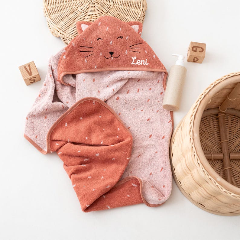 May include: A coral and pink hooded baby towel with a cat face design. The towel has white polka dots and the name "Leni" embroidered on the hood. A matching washcloth is folded next to the towel. A bottle of lotion and wooden blocks are also in the image.