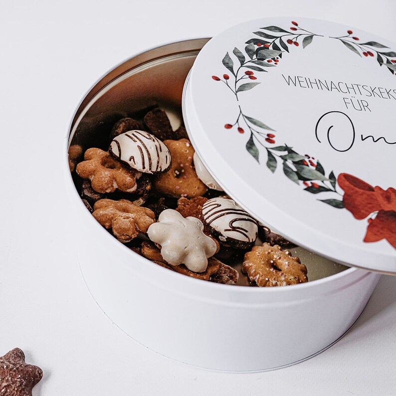 May include: A white, round tin filled with assorted biscuits. The lid, slightly ajar, features a wreath design with red berries and the text "WEIHNACHTSKEKS F&Uuml;R Oma". The biscuits are various shapes and colours, some with white icing.