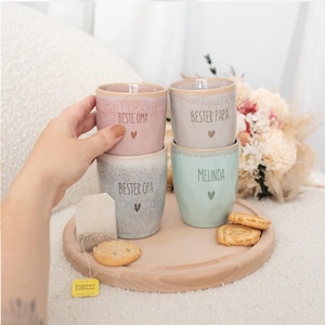 Puede incluir: Cuatro tazas de cer&aacute;mica en colores pastel, apiladas sobre una bandeja de madera. Cada taza tiene texto y un s&iacute;mbolo de coraz&oacute;n. Las tazas son rosa, gris, azul claro y beige. Una bolsita de t&eacute; y galletas tambi&eacute;n est&aacute;n en la bandeja.