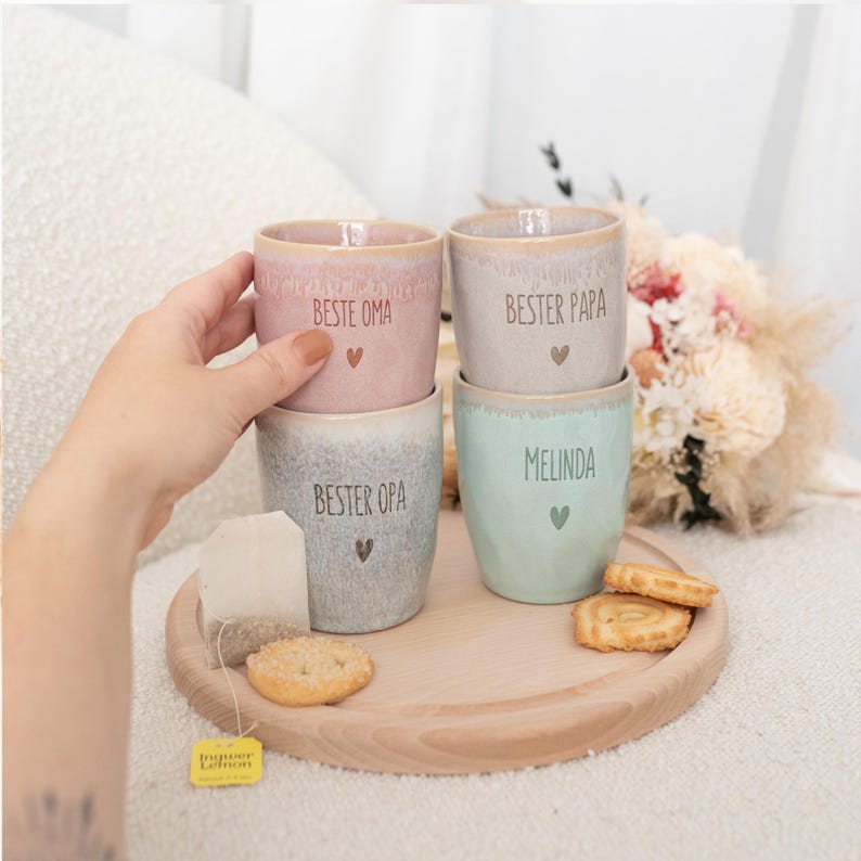 Puede incluir: Cuatro tazas de cer&aacute;mica en colores pastel, apiladas sobre una bandeja de madera. Cada taza tiene texto y un s&iacute;mbolo de coraz&oacute;n. Las tazas son rosa, gris, azul claro y beige. Una bolsita de t&eacute; y galletas tambi&eacute;n est&aacute;n en la bandeja.