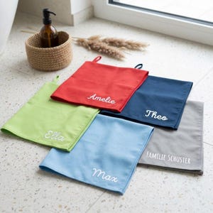 Può includere: Una collezione di asciugamani colorati e personalizzati. Gli asciugamani sono nei toni del rosso, verde, blu, blu navy e grigio. Ogni asciugamano ha un nome ricamato in bianco. Un cestino intrecciato e una bottiglia marrone sono sullo sfondo.