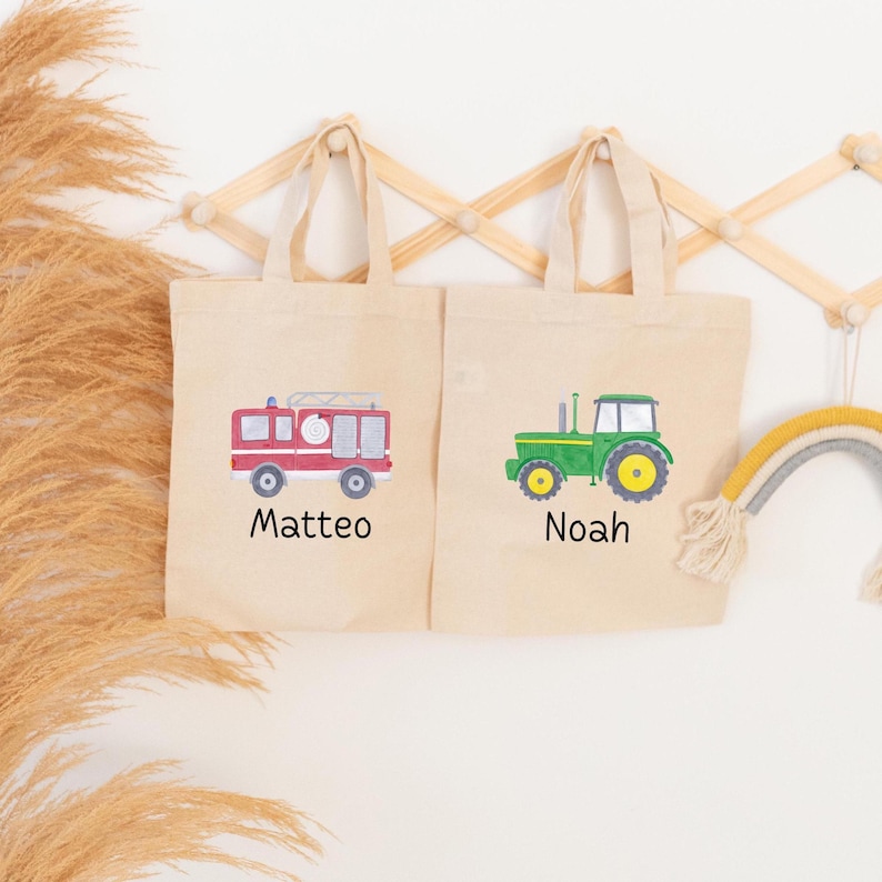 May include: Two beige canvas tote bags with handles. The left bag features a red fire truck graphic with the name Matteo printed below. The right bag features a green tractor graphic with the name Noah printed below in black lettering.