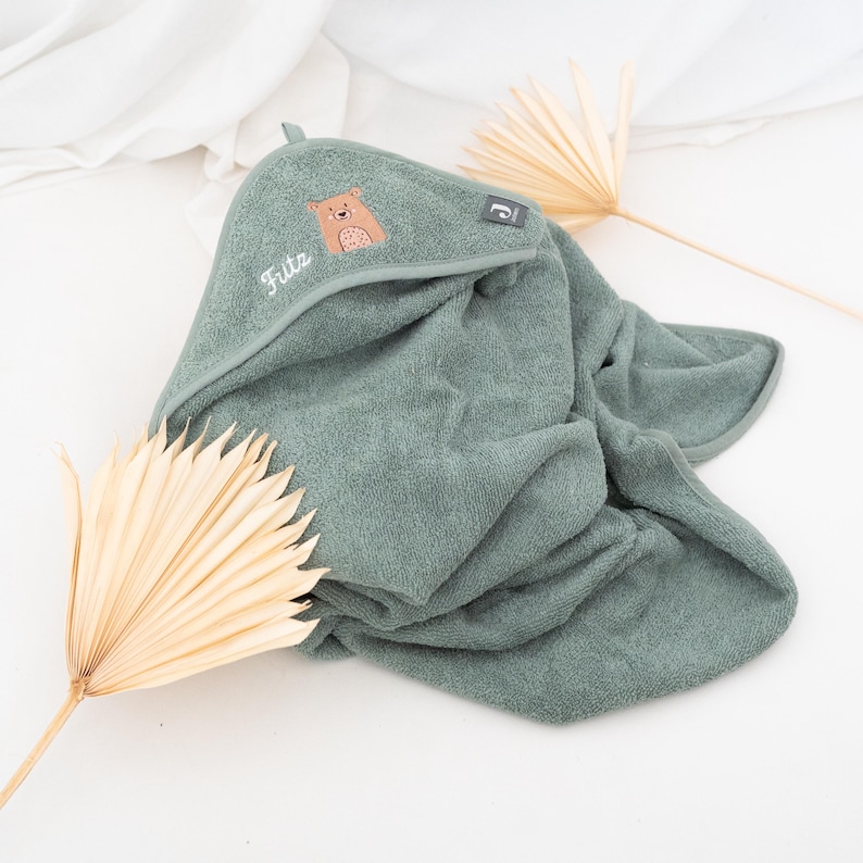 May include: A sage green hooded baby towel with a bear applique and the name "Fritz" embroidered in white. The towel is made of soft terry cloth and has a small loop for hanging. A small tag with a logo is visible on the hood.