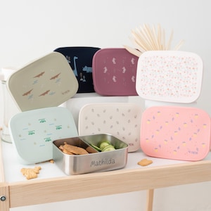 Op de afbeelding: Een verzameling kleurrijke rechthoekige lunchboxen met diverse ontwerpen, waaronder dinosaurussen, zeepaardjes en bloempatronen. Eén doos is open en toont een roestvrijstalen interieur met voedsel en de naam "Mathilda" gegraveerd.