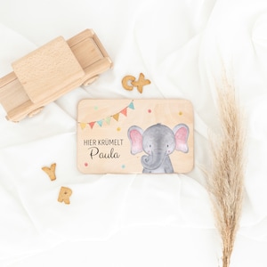 May include: A rectangular wooden board featuring a watercolor illustration of a gray elephant with pink ears, colorful bunting, and the text HIER KR&Uuml;MELT Paula. The board is accompanied by a wooden toy truck and several small letter-shaped cookies.