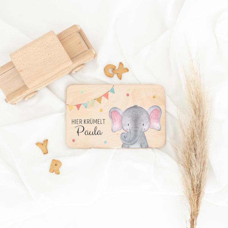 May include: A rectangular wooden board featuring a watercolor illustration of a gray elephant with pink ears, colorful bunting, and the text HIER KR&Uuml;MELT Paula. The board is accompanied by a wooden toy truck and several small letter-shaped cookies.