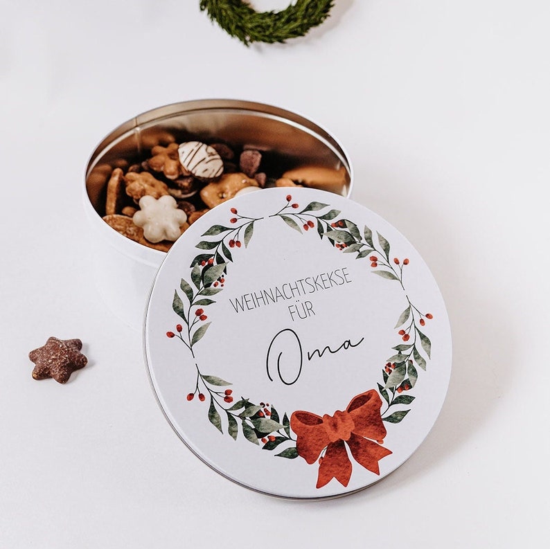 May include: A white, round tin filled with assorted biscuits, with the lid slightly ajar. The lid features a watercolour wreath design with red berries and a red bow, and the text "WEIHNACHTSKEKSE F&Uuml;R Oma". A star-shaped biscuit sits nearby.