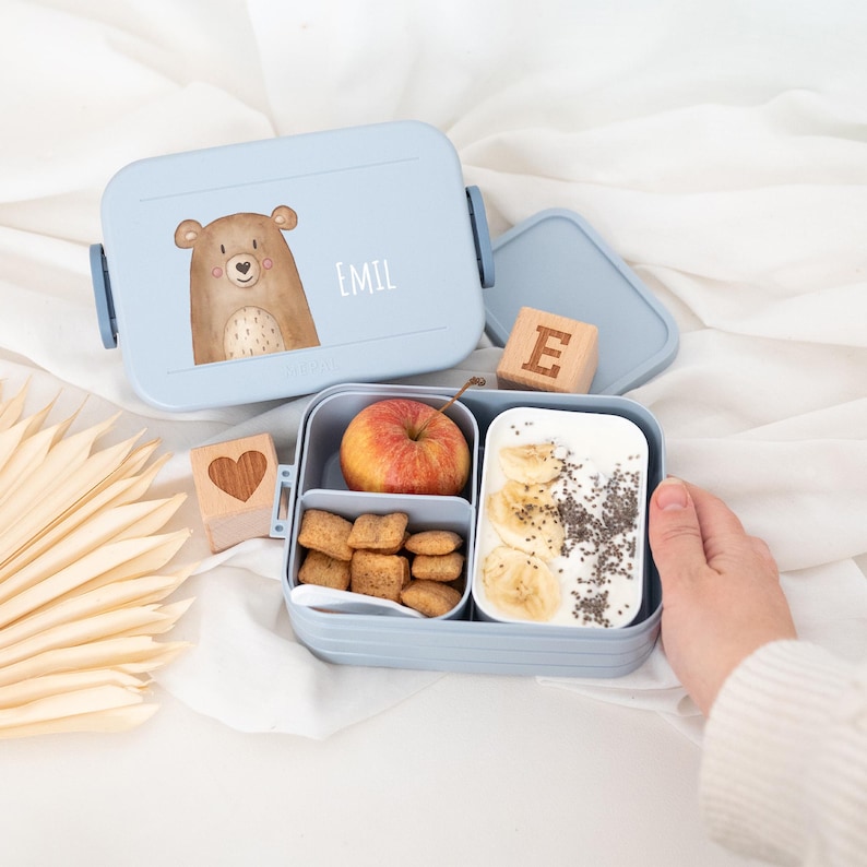 May include: A blue lunchbox with a bear illustration and the name "EMIL" on the lid. The lunchbox is open and contains an apple, crackers, and yogurt with banana and chia seeds. There are also two wooden blocks, one with a heart and the other with the letter "E".