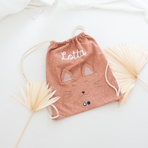 May include: A brown drawstring bag with a cat face design and the name "Lotta" embroidered on it. The bag has white stitching and black eyes.