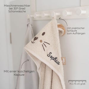 May include: A beige hooded baby towel with embroidered facial features and the name "Sophia". The towel has a loop for hanging and measures 75 x 75 cm.