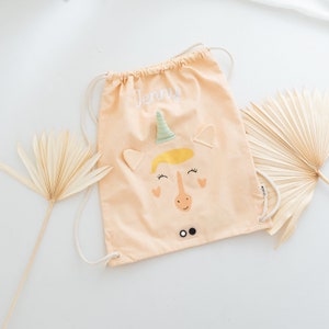 May include: A light peach drawstring bag with a unicorn design. The unicorn has a yellow mane, green horn, and black eyes. The bag has the name "Jenny" embroidered on it.
