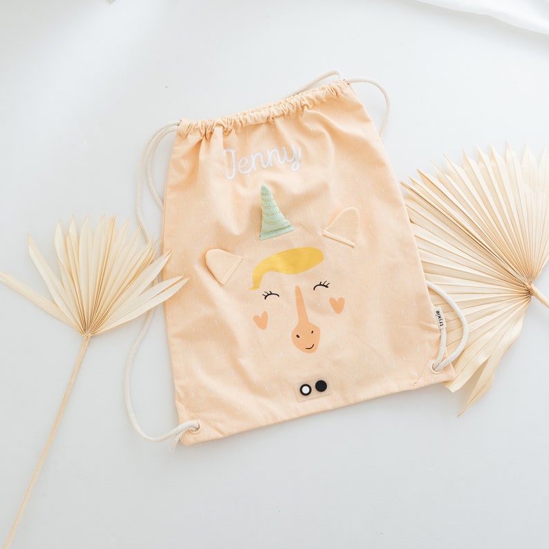 May include: A light peach drawstring bag with a unicorn design. The unicorn has a yellow mane, green horn, and black eyes. The bag has the name "Jenny" embroidered on it.