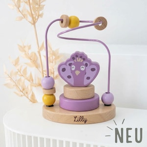 Peut inclure: Un jouet labyrinthe de perles en bois avec un motif de paon dans des tons de violet, jaune et bois naturel. Le jouet comprend une piste m&eacute;tallique avec des perles et une base grav&eacute;e du nom "Lilly". Le mot "NEU" est &eacute;galement visible.