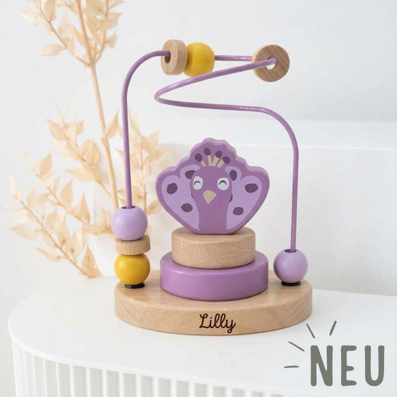 Peut inclure: Un jouet labyrinthe de perles en bois avec un motif de paon dans des tons de violet, jaune et bois naturel. Le jouet comprend une piste m&eacute;tallique avec des perles et une base grav&eacute;e du nom "Lilly". Le mot "NEU" est &eacute;galement visible.