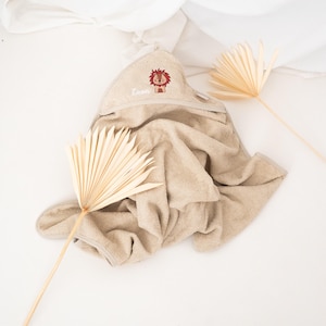 May include: A beige hooded baby towel with a lion design embroidered on the hood. The lion is brown and red, and the name "Leon" is embroidered in white. The towel is made of soft material and is folded on a white surface.