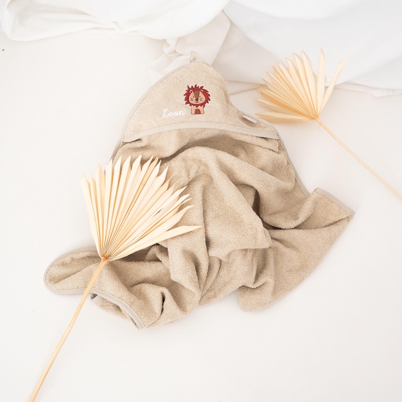 May include: A beige hooded baby towel with a lion design embroidered on the hood. The lion is brown and red, and the name "Leon" is embroidered in white. The towel is made of soft material and is folded on a white surface.