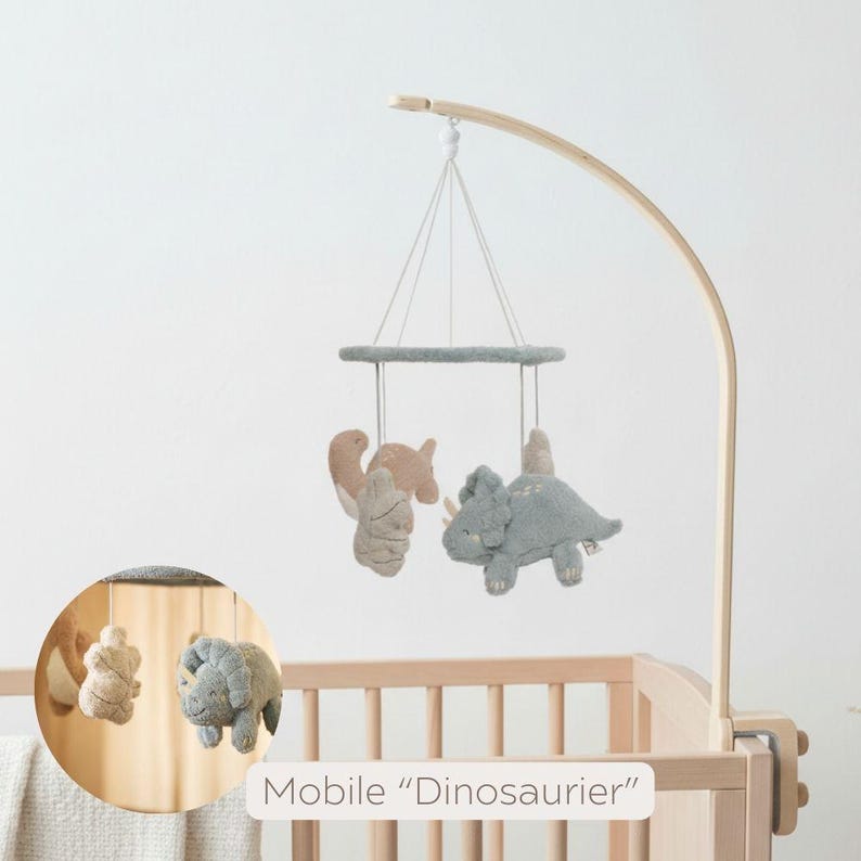May include: A baby mobile featuring three plush dinosaur figures in beige and gray tones suspended from a gray ring. The mobile is attached to a wooden curved arm, and the text "Mobile 'Dinosaurier'" is visible.