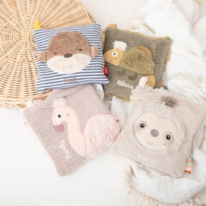 Può includere: Quattro giocattoli di peluche per bambini con facce di animali: una lontra, un cammello, un fenicottero e un bradipo. La lontra è su un cuscino a righe blu e bianche. Il cammello è su un cuscino verde. Il fenicottero è su un cuscino rosa. Il bradipo è su un cuscino beige.