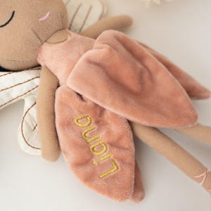 May include: A plush fairy doll in soft, muted tones. The doll features a tan face and limbs, a pink dress, and matching wings. The wings are embroidered with the name "Liana" in gold thread. The doll's ballet slippers have pink ribbons.