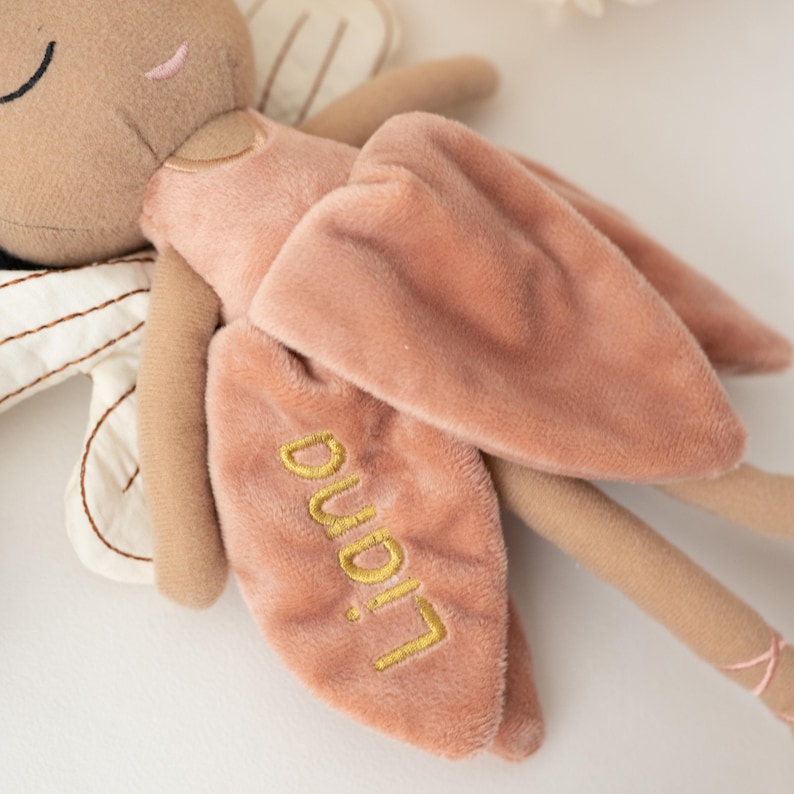 May include: A plush fairy doll in soft, muted tones. The doll features a tan face and limbs, a pink dress, and matching wings. The wings are embroidered with the name "Liana" in gold thread. The doll's ballet slippers have pink ribbons.