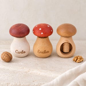 Op de afbeelding: Drie houten objecten in de vorm van een paddenstoel. De eerste is van licht hout met een donkerrode hoed en de naam Carlo gegraveerd. De tweede is van licht hout met een rode hoed met witte stippen en de naam Emilia gegraveerd. De derde is een notenkraker.