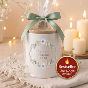 Op de afbeelding: Een witte keramische pot met een houten deksel, verpakt in cellofaan en vastgebonden met een saliegroen lint. De pot heeft een bloemenkransontwerp met tekst. Een "Bestseller"-sticker is zichtbaar.