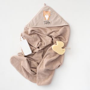 May include: A beige hooded towel with a cartoon cat and the name "Tilda" embroidered on the hood. A white bottle of lotion and a yellow rubber duck are also pictured.