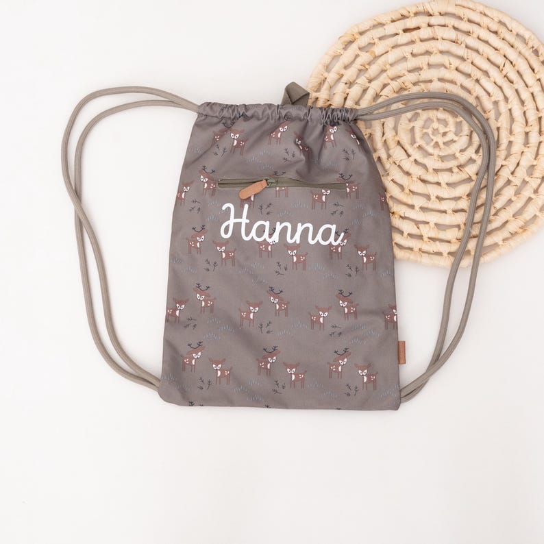 May include: A gray drawstring backpack with a repeating pattern of brown deer and the name "Hanna" in white script. The bag has a small zippered pocket and gray drawstrings. A woven circular placemat is in the background.