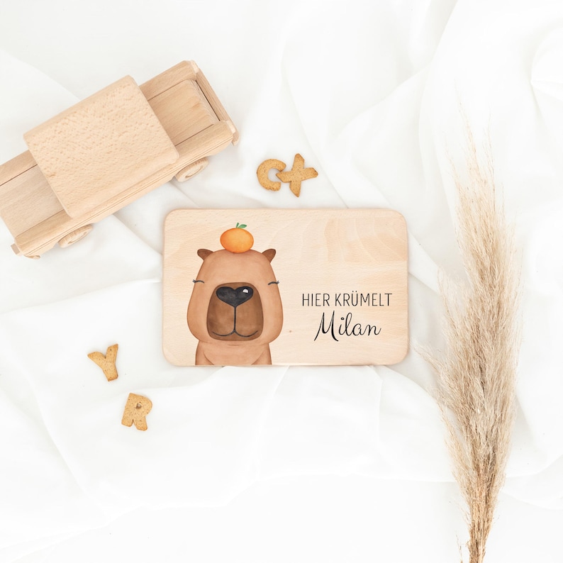 May include: A rectangular wooden board featuring a brown capybara illustration with an orange on its head and the text "HIER KR&Uuml;MELT Milan" in black. The board is accompanied by a wooden toy truck and small wooden letter shapes.