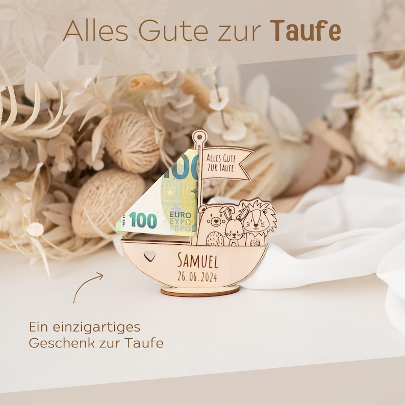 May include: A wooden sailboat with a &euro;100 note inside. The sailboat has a heart-shaped cutout and the text "Alles Gute zur Taufe" on the sail. The sailboat also has the text "SAMUEL 26.06.2024" on the bottom. The sailboat is surrounded by dried flowers.