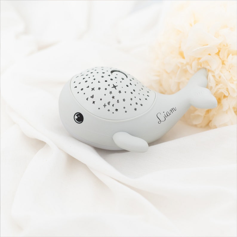 May include: A light gray whale-shaped night light featuring a perforated top surface with star and moon cutouts. The side of the whale is personalized with the name Liam in a black cursive script, and it has a simple black eye graphic.
