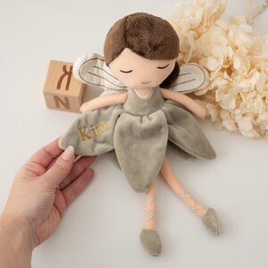 May include: A soft, plush fairy doll in muted green with brown hair and closed eyes. The doll has embroidered wings and a dress, with the name "Kim" embroidered in gold. The doll's legs are light pink with ballet-style shoes.