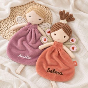 Può includere: Due bambole fata in peluche con nomi ricamati. La bambola a sinistra ha capelli biondi, un vestito color malva con il nome Amelie e ali semplici. Quella a destra ha capelli castani, un vestito color terracotta con il nome Selma e ali con dettagli a cuore.