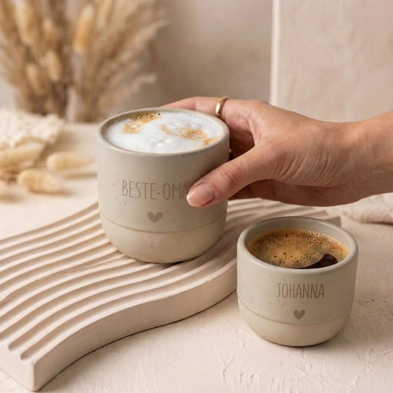 Pode incluir: Duas canecas de cer&acirc;mica bege com acabamento salpicado. A caneca maior cont&eacute;m um latte espumoso e est&aacute; inscrita com o texto 'BESTE OMA' acima de um pequeno cora&ccedil;&atilde;o. A caneca menor cont&eacute;m caf&eacute; preto e o nome 'JOHANNA' acima de um cora&ccedil;&atilde;o.