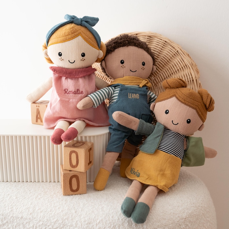 May include: Three handmade fabric dolls with embroidered faces. One doll wears a pink dress with the name "Rosalie", another wears blue overalls with the name "Leano", and the third wears a yellow skirt with the name "Olivia". Each doll has a unique hairstyle and outfit.