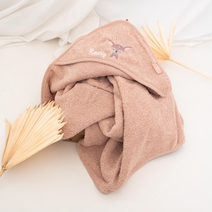May include: A dusty rose hooded baby towel. The towel features an embroidered deer design and the name "Romy" in script. The towel is folded, with the hood visible, and is made of a soft, absorbent material. A dried palm leaf is in the background.