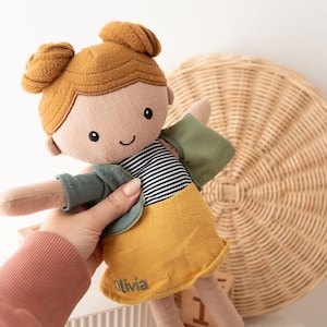 May include: A handmade doll with a smiling face, brown hair in two buns, and a striped shirt. The doll wears a green jacket and a yellow skirt. The name "Olivia" is embroidered on the skirt. The doll is being held in a person's hand.
