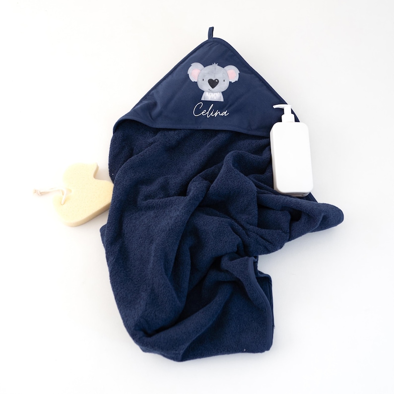 May include: A navy blue hooded towel with a koala design and the name "Celina" embroidered on the hood. A white bottle of lotion and a yellow sponge are also in the image.